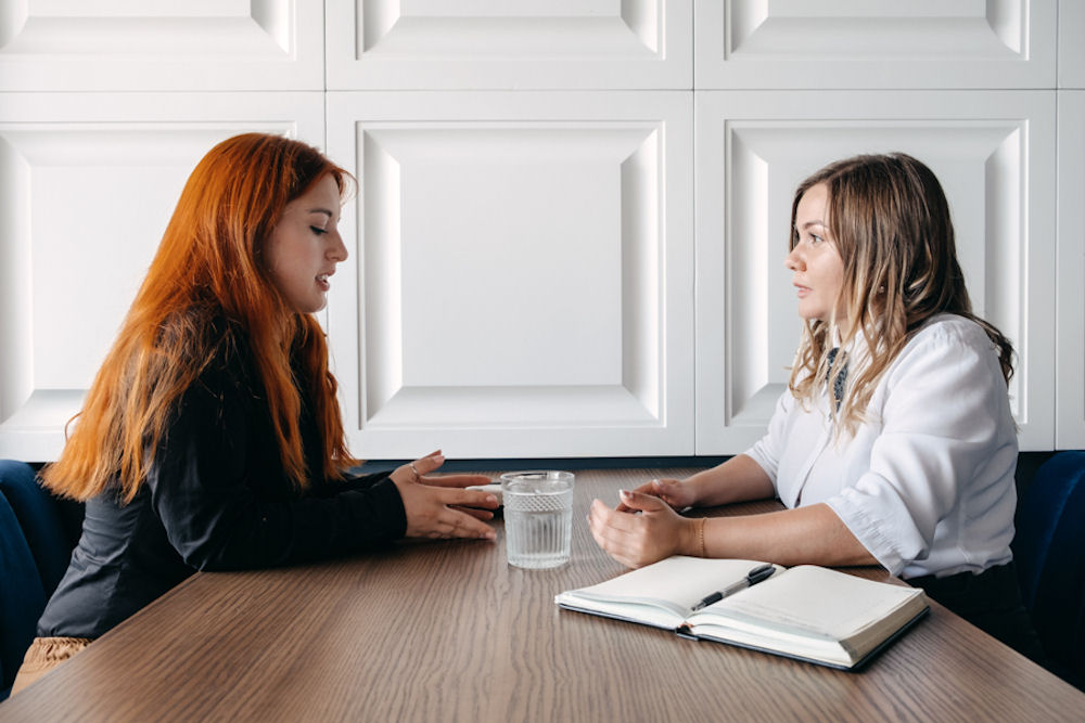 Young woman with red hair talking openly with older blind woman at long table