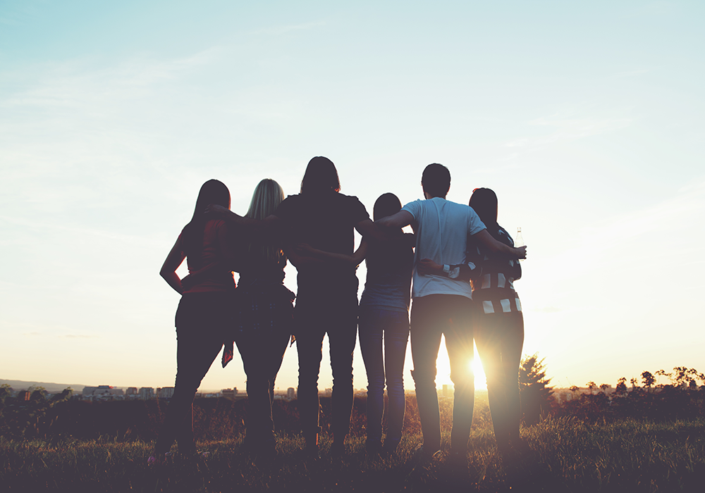 Rear view of four women and two men with arms around each other, facing the sunset