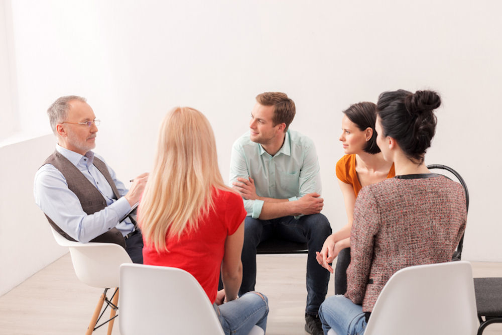 Sitting addiction treatment group looking at facilitator with notepad