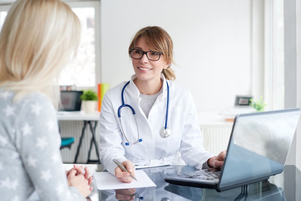 Doctor with glasses taking notes on laptop and notepad as woman across from her speaks