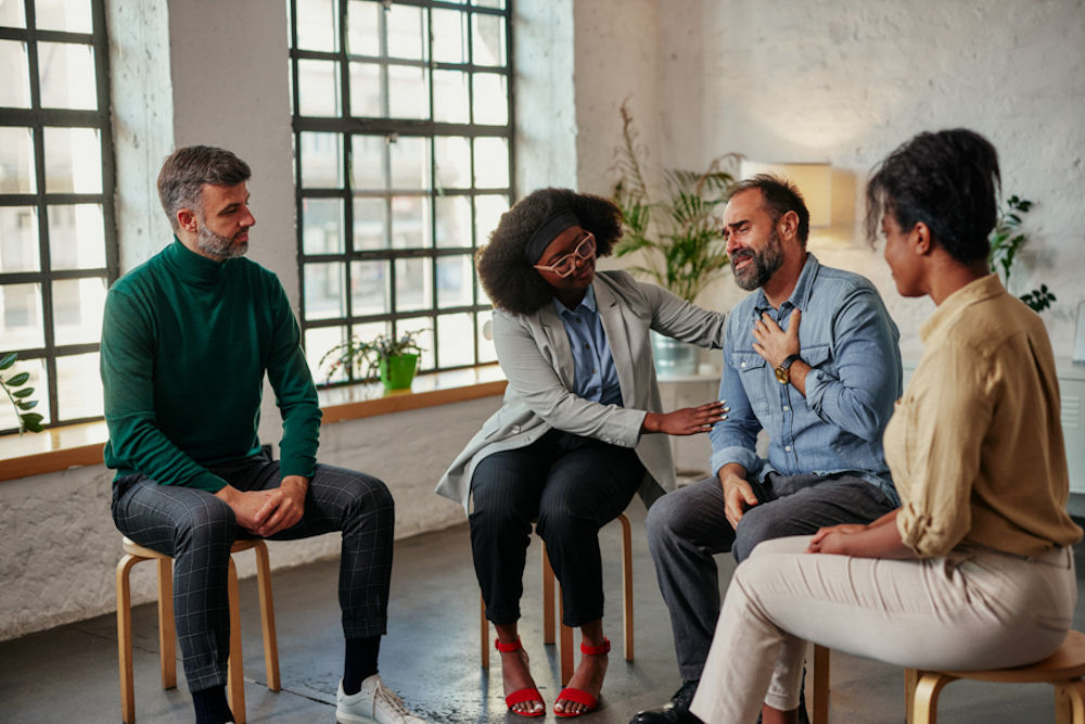 Young woman consoling older man in group therapy session with large windows and potted plants