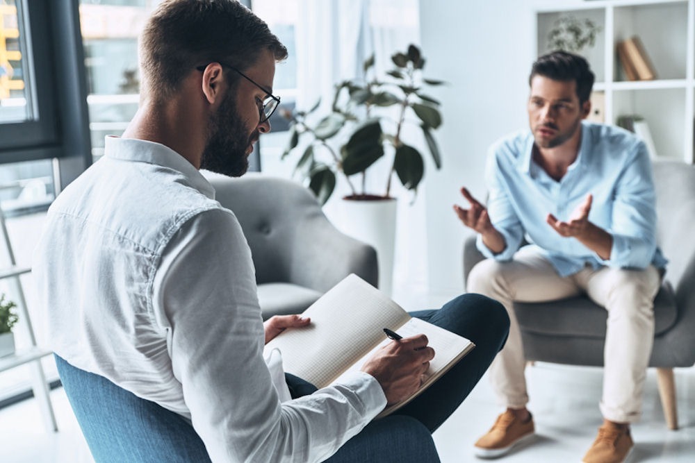 Seated man explaining things to male therapist taking notes in notebook