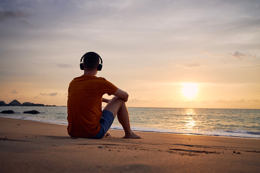 Man wearing cans headphones thinking about a sober new year while sitting on a sunset beach